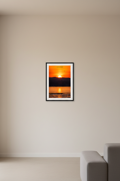 Small fine art portrait print of a fiery telephoto sunrise with massive sun rising above silhouetted treeline over partially frozen Lake Leelanau with golden reflection path and single bird in flight February 27 2024 displayed in modern living room