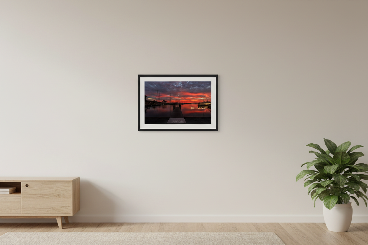 Small fine art print of blazing red fire sunrise over Northport Marina with dock leading line sailboat mast silhouettes and perfect mirror reflection in calm harbor water displayed in modern living room