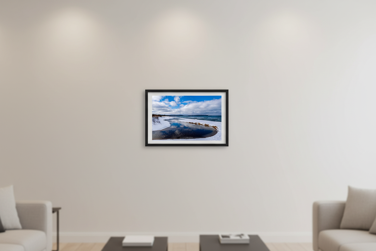 Small fine art landscape print of curved tidal pool reflecting blue sky and clouds on snow-covered Good Harbor Bay shoreline with turquoise Lake Michigan and Sleeping Bear Bluffs on horizon January 29 2023 displayed in modern living room