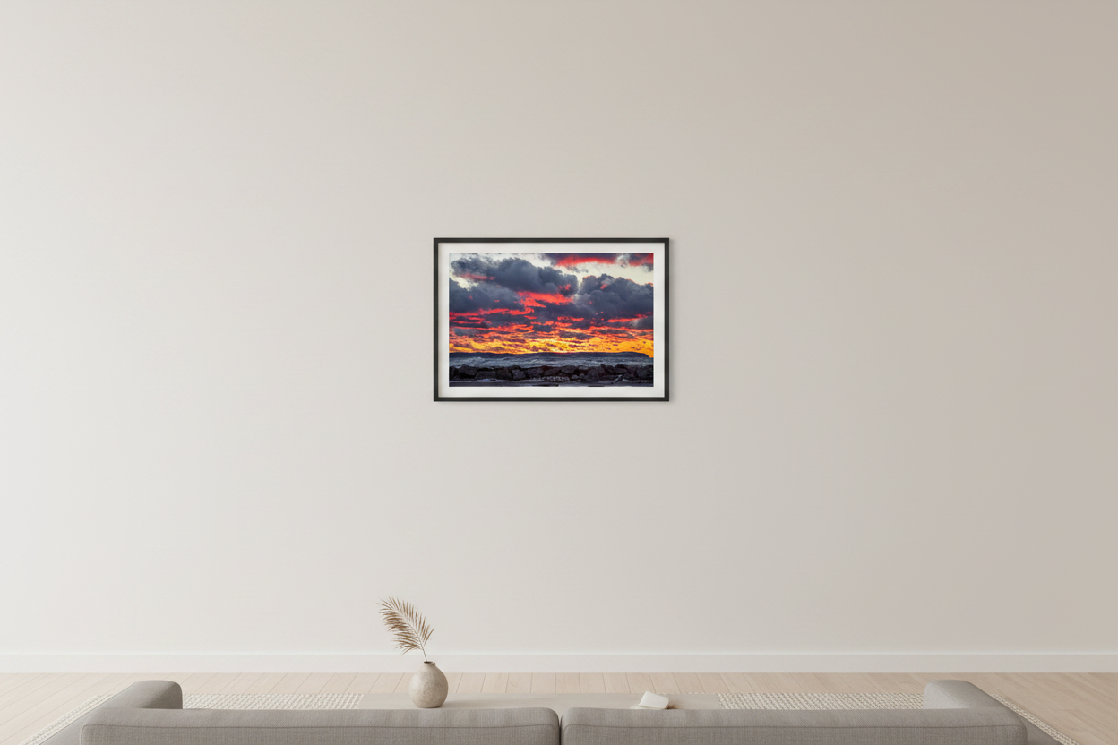 Small fine art landscape print of explosive fiery crimson and magenta layered clouds lit like flames over golden-orange horizon with crashing white-capped waves against rocky breakwall and dark distant headland silhouette at Leland Lake Michigan December 2021 displayed in modern living room