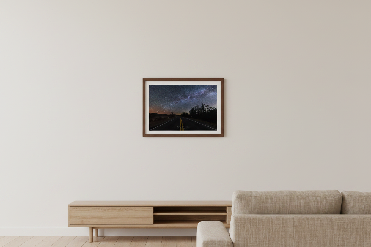 Small fine art landscape print of the Milky Way arch over a two-lane highway with double yellow center lines vanishing into the distance at Sleeping Bear Dunes Michigan April 7 2024 at 4:17am with warm amber horizon glow and silhouetted tree line displayed in modern living room