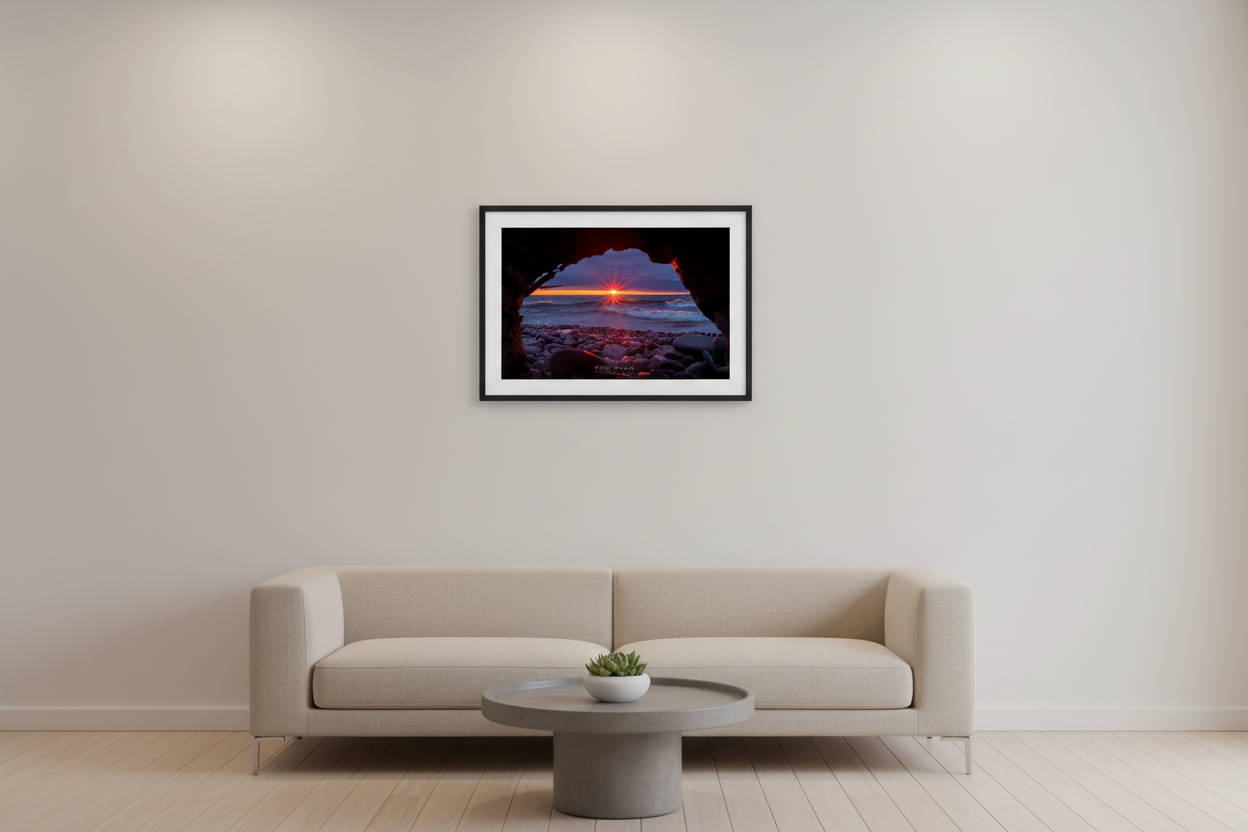 Small fine art landscape print of starburst sunset framed through driftwood rock arch over crashing waves on smooth pebble beach at Eagle River Lake Superior Michigan April 2023 displayed in modern living room