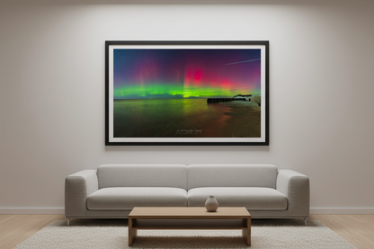 Large fine art landscape print of explosive full-spectrum Northern Lights with vivid lime green base erupting into tall crimson red and magenta pillars filling the sky reflecting green on calm water with sandy beach and dark wooden pier at Point Betsie Lake Michigan Michigan February 26 2023 displayed in modern living room