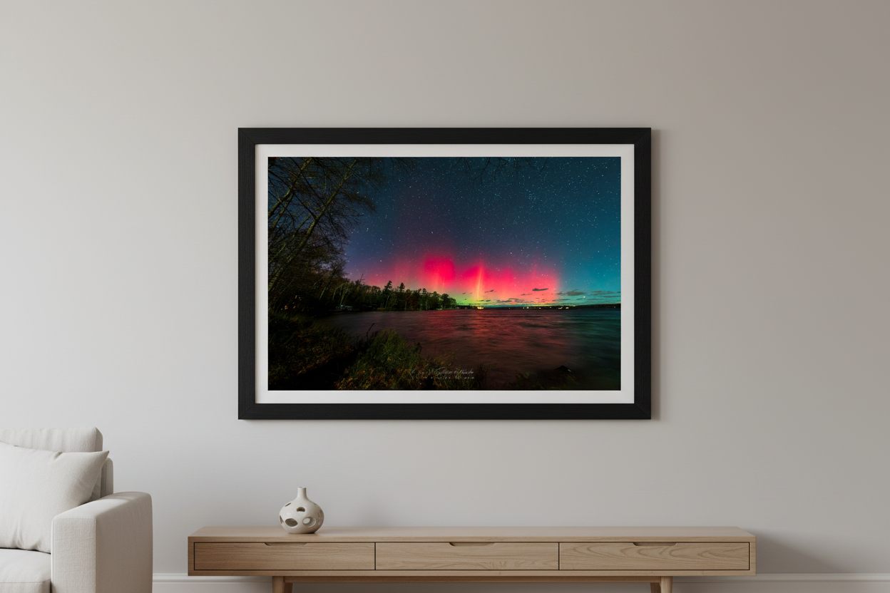 Large fine art print of double-lobe crimson red aurora arches glowing over Lake Leelanau wooded shoreline from Leland Michigan November 8 2024 8:27pm with pink water reflection and starfield displayed in modern living room