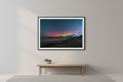 Large fine art landscape print of vivid Northern Lights with magenta purple and neon green aurora bands over glassy calm Lake Michigan shoreline with dark forested peninsula silhouetted on horizon and sandy beach at Fisherman's Island Charlevoix Michigan August 19 2022 displayed in modern living room