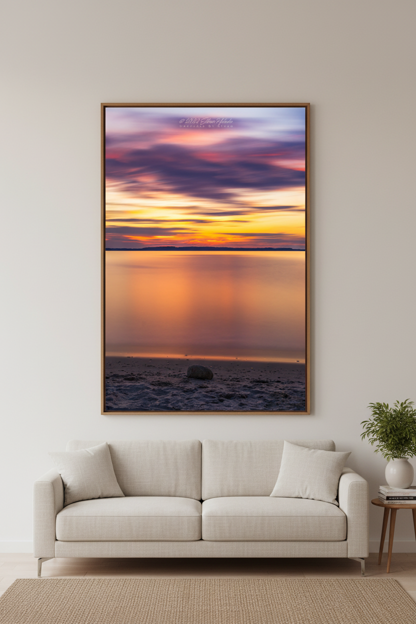 Large fine art portrait print of 6-minute long exposure sunset with silky smooth mirror-like water reflecting blazing orange and gold horizon with streaking pink and purple clouds and single smooth stone on sandy beach at Elk Rapids Grand Traverse Bay Michigan August 2022 displayed in modern living room