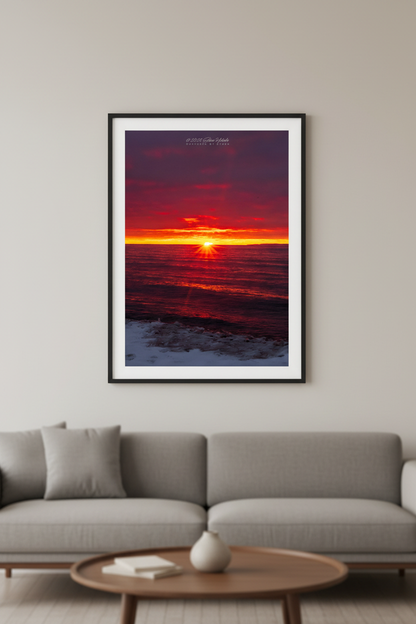 Large fine art portrait print of starburst sunset with deep crimson and purple clouds blazing yellow-orange horizon golden reflection across dark Lake Michigan waves and snow-dusted icy shoreline at Van's Beach Leland Michigan March 2023 displayed in modern living room