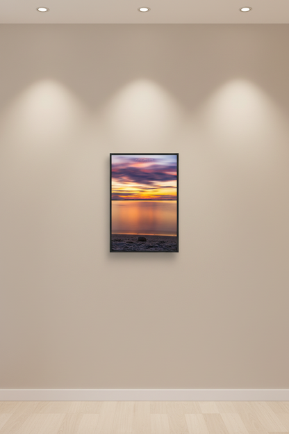 Small fine art portrait print of 6-minute long exposure sunset with silky smooth mirror-like water reflecting blazing orange and gold horizon with streaking pink and purple clouds and single smooth stone on sandy beach at Elk Rapids Grand Traverse Bay Michigan August 2022 displayed in modern living room