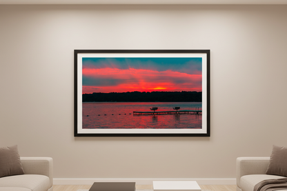 Large fine art print of Lake Leelanau fire sunset with visible golden sun orb at treeline centered L-shaped swimming dock with benches magenta pink sky teal storm clouds wide open water foreground swim rope buoy line Leland Michigan displayed in modern living room