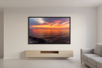 Large fine art landscape print of a fiery orange and magenta sunset over calm Lake Michigan waters with rocky shoreline foreground viewed from Leland Michigan July 2024 displayed in modern living room