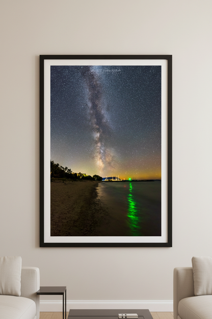 Large fine art portrait print of Milky Way rising over calm lake shoreline with vivid green navigation light reflecting in water sailboats and marina lights glowing in distance and sandy beach curving in foreground at Fishtown Leland Michigan August 21 2022 displayed in modern living room