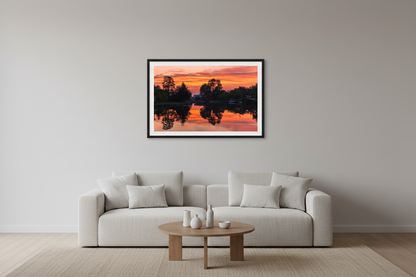 Small fine art landscape print of perfectly mirrored blazing orange pink and crimson sunset sky reflected in glassy calm Leland River channel with dark tree silhouettes framing both sides and docks along the banks July 1 2023 displayed in modern living room