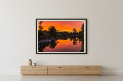 Large fine art landscape print of blazing orange red sunset perfectly mirrored in glassy Leland River with boat docks and tree silhouettes Michigan September 2022 displayed in modern living room