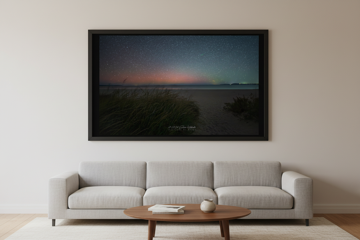 Large fine art print of aurora borealis pink red glow over Bohemian Road Beach Michigan dune grass clumps open sandy beach calm dark water distant landmass starfield sky Oct 5 2024 displayed in modern living room