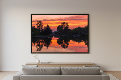 Large fine art landscape print of perfectly mirrored blazing orange pink and crimson sunset sky reflected in glassy calm Leland River channel with dark tree silhouettes framing both sides and docks along the banks July 1 2023 displayed in modern living room