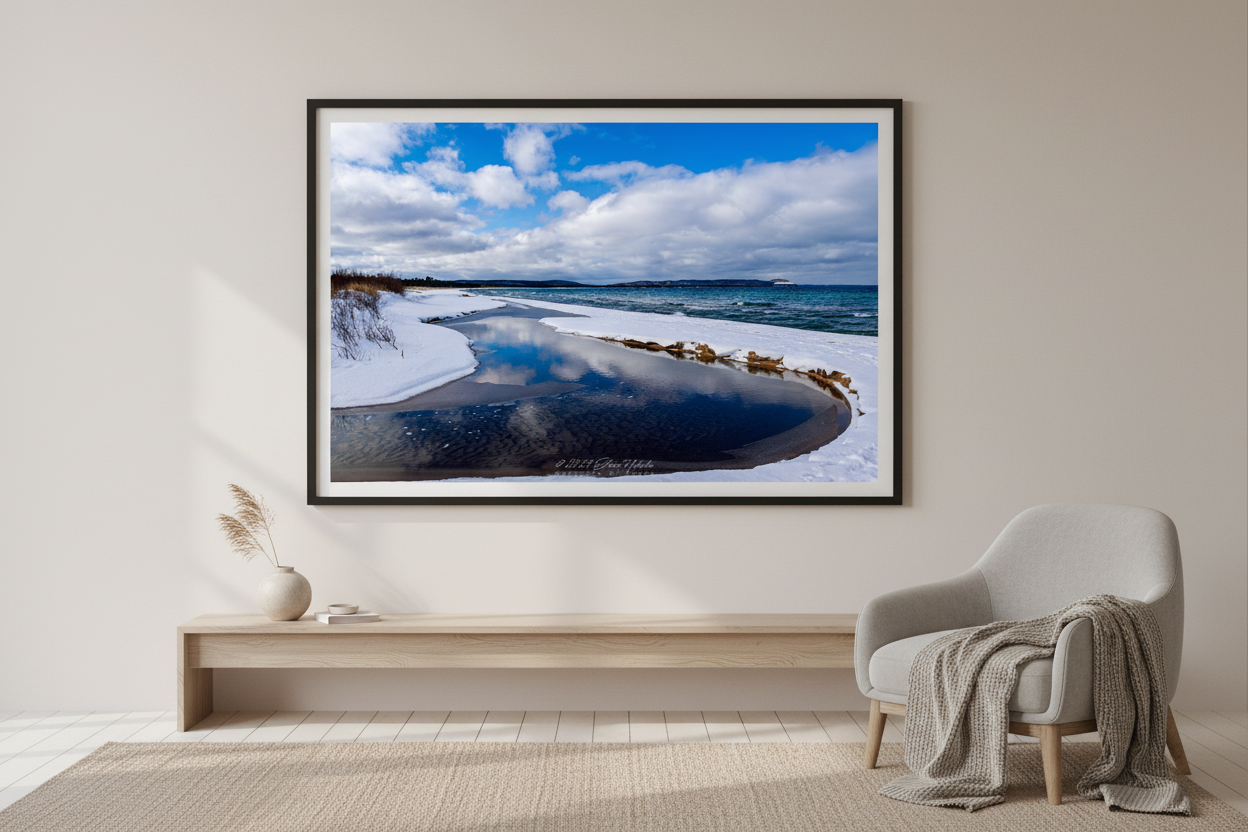 Large fine art landscape print of curved tidal pool reflecting blue sky and clouds on snow-covered Good Harbor Bay shoreline with turquoise Lake Michigan and Sleeping Bear Bluffs on horizon January 29 2023 displayed in modern living room