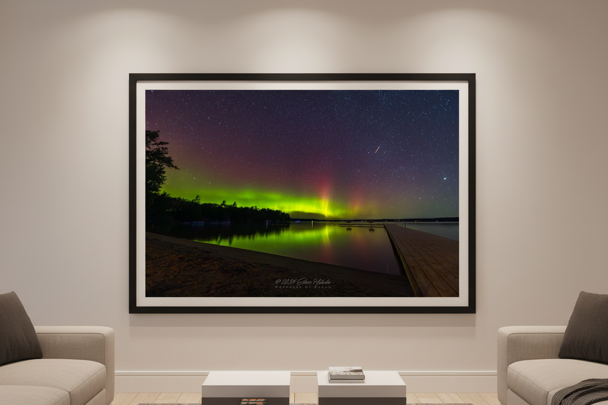 Large fine art print of vertical aurora pillars lime green and crimson over Lake Leelanau with wooden dock foreground deciduous treeline and glassy lake reflection meteor streak Leland Michigan displayed in modern living room