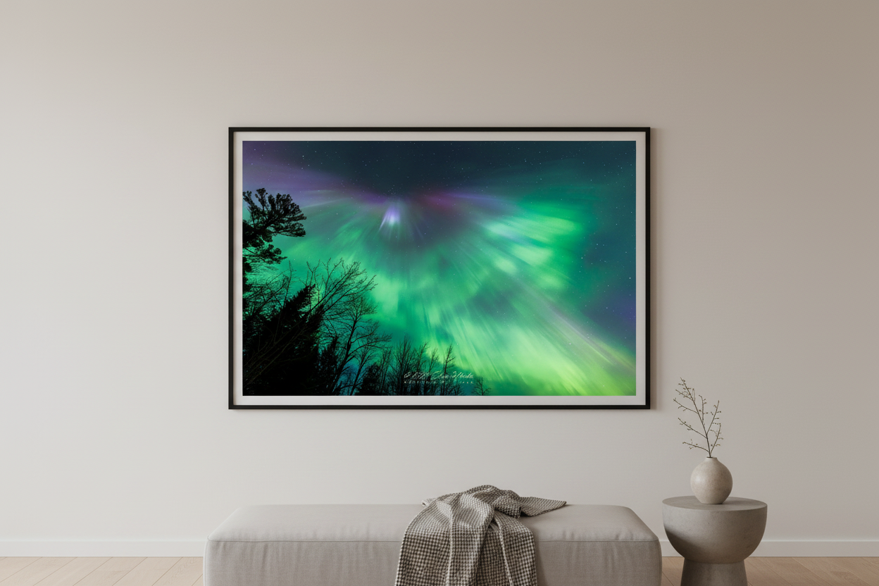Large fine art landscape print of vivid green aurora corona burst with purple magenta corona point radiating curtains above silhouetted trees Lake Superior shoreline Michigan Upper Peninsula April 24 2023 displayed in modern living room