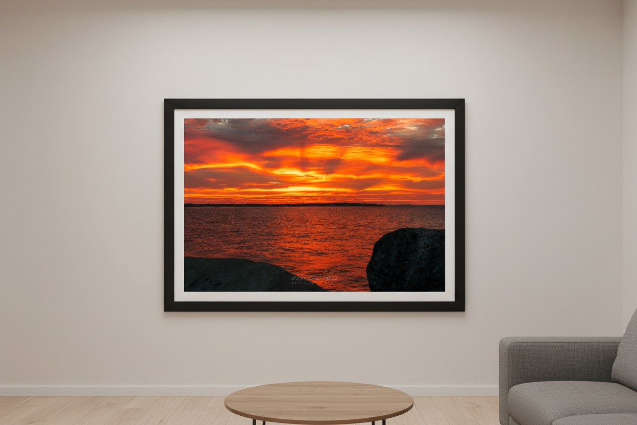 Large fine art print of blazing orange amber fire sunrise over Northport Marina with rocky foreground island silhouette and warm glowing water displayed in modern living room