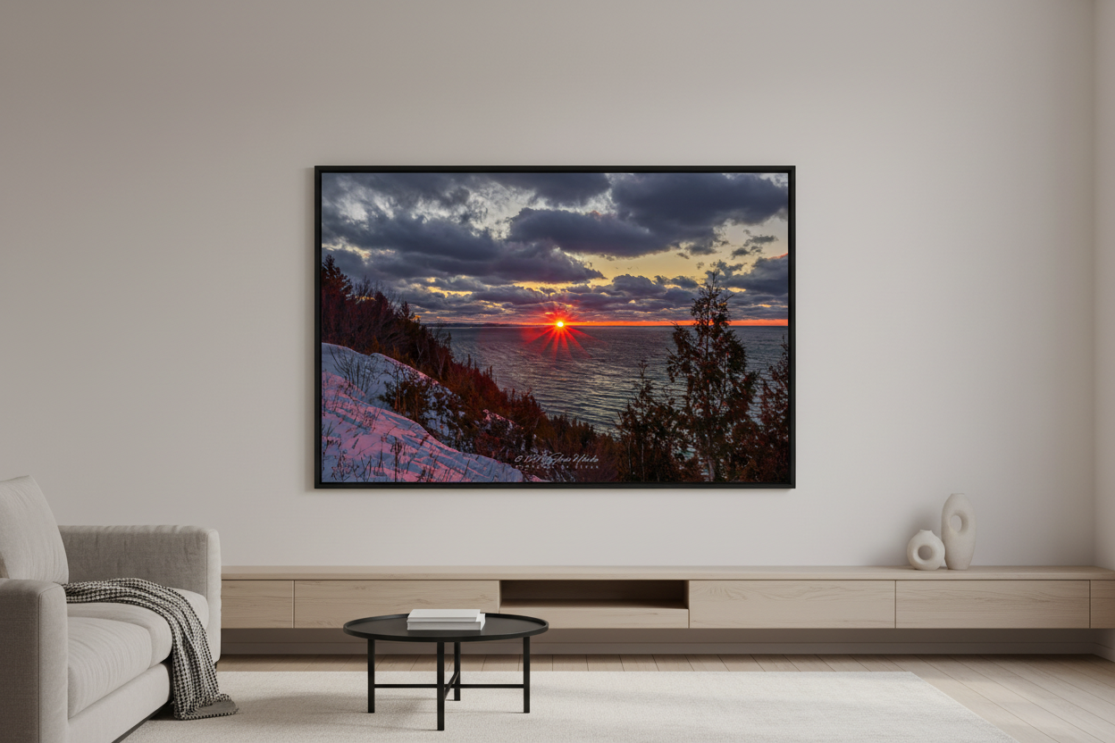 Large fine art landscape print of dramatic starburst sunset with fiery red sun touching Lake Michigan horizon under moody dark storm clouds with golden edges viewed from snow-covered bluff with silhouetted evergreen trees at Sleeping Bear Dunes January 2023 displayed in modern living room