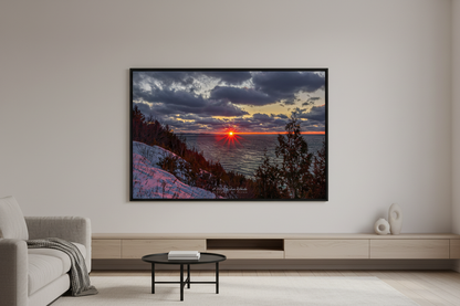 Large fine art landscape print of dramatic starburst sunset with fiery red sun touching Lake Michigan horizon under moody dark storm clouds with golden edges viewed from snow-covered bluff with silhouetted evergreen trees at Sleeping Bear Dunes January 2023 displayed in modern living room
