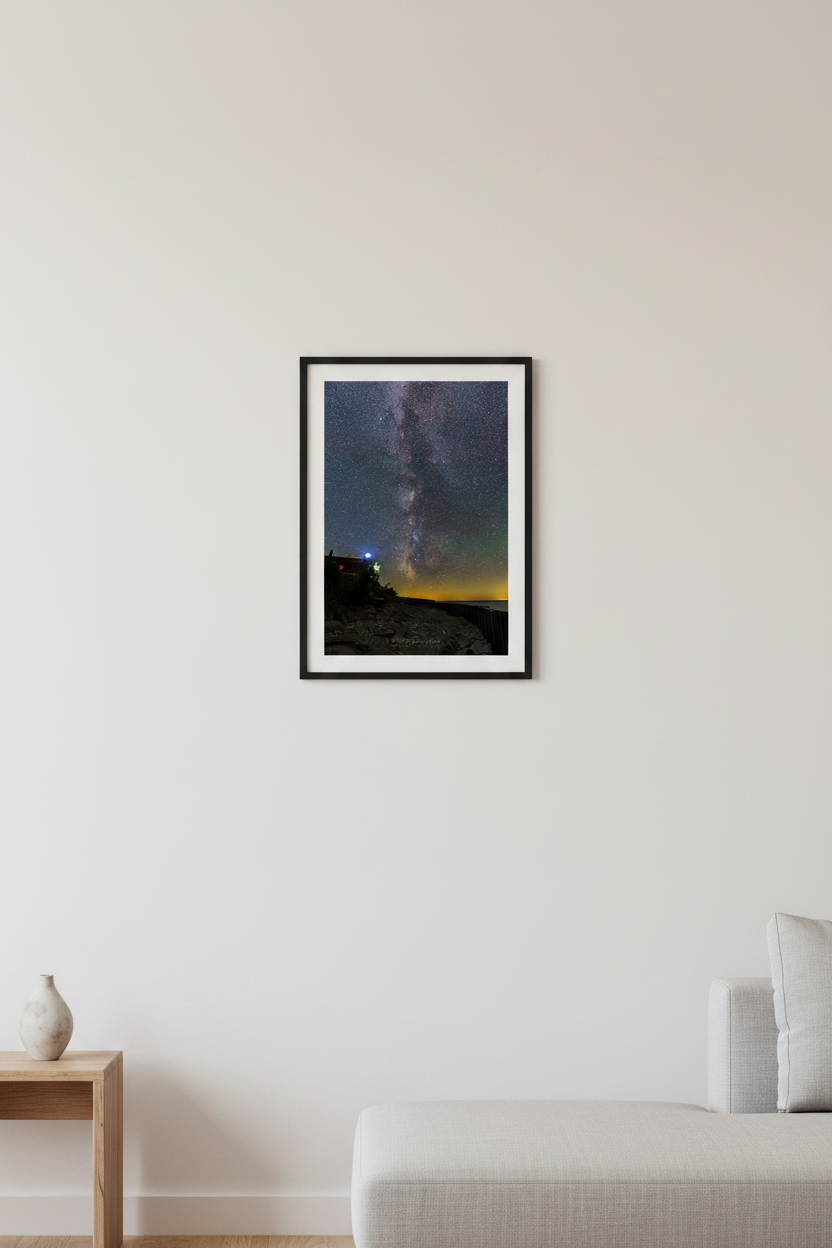 Small fine art portrait print of the Milky Way galactic core rising above the glowing Point Betsie Lighthouse on Lake Michigan Michigan August 5 2022 at 1:32am with amber horizon glow and rocky shoreline displayed in modern living room