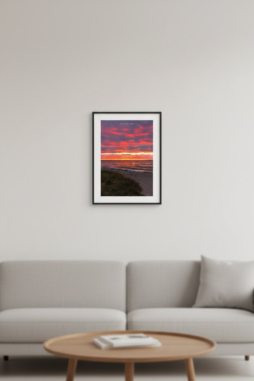 Small fine art portrait print of fiery red and magenta cloud explosion over Lake Michigan with dune grass foreground and gentle shoreline waves at Van's Beach Leland MI October 2023 displayed in modern living room
