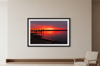 Large fine art print of fiery crimson red orange sunrise over Suttons Bay Leelanau County Michigan with wooden dock pilings silhouette dark treeline reflection in calm water November 3 2024 displayed in modern living room