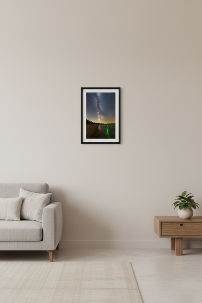 Small fine art portrait print of Milky Way rising over calm lake shoreline with vivid green navigation light reflecting in water sailboats and marina lights glowing in distance and sandy beach curving in foreground at Fishtown Leland Michigan August 21 2022 displayed in modern living room