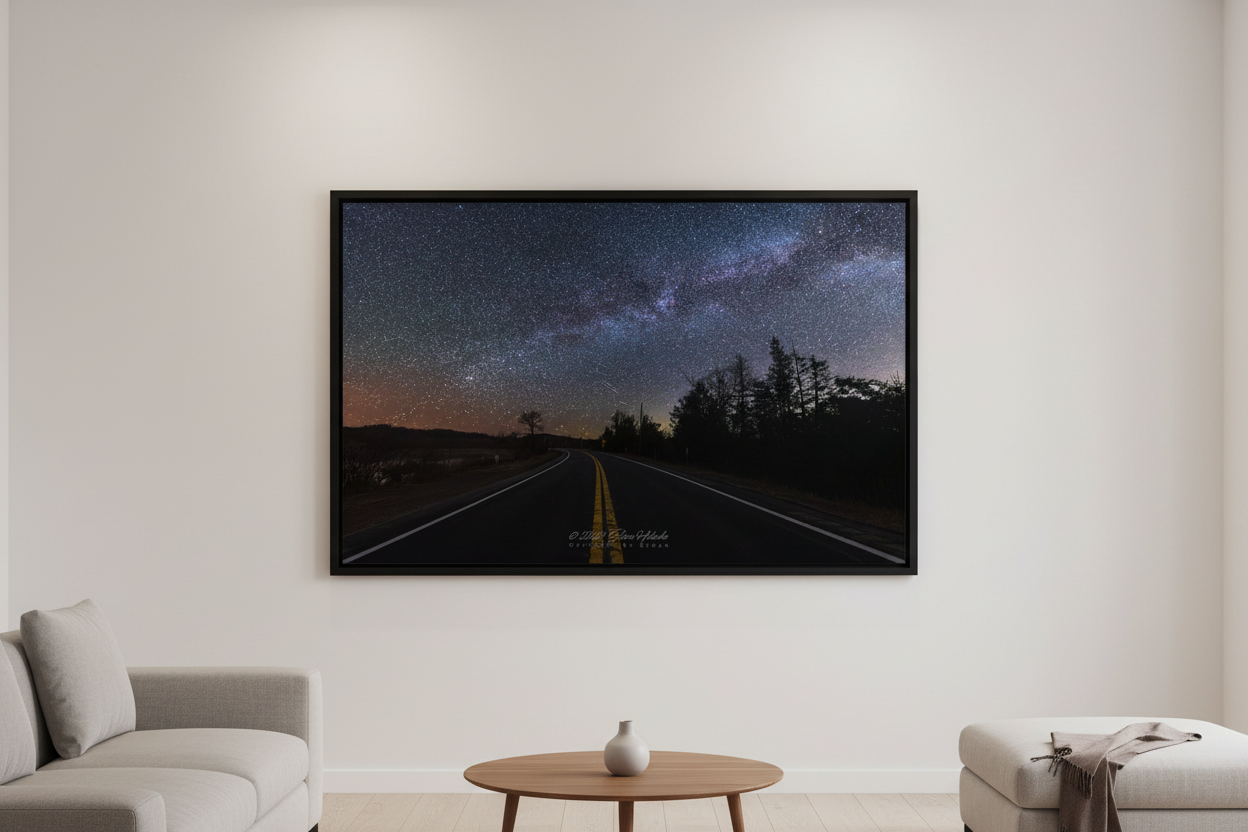 Large fine art landscape print of the Milky Way arch over a two-lane highway with double yellow center lines vanishing into the distance at Sleeping Bear Dunes Michigan April 7 2024 at 4:17am with warm amber horizon glow and silhouetted tree line displayed in modern living room