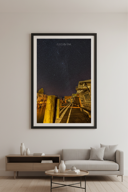 Large fine art portrait print of star-filled night sky over Fishtown docks Leland River with warm golden dock lighting and DAM Candy Store Leland Michigan February 11 2023 displayed in modern living room