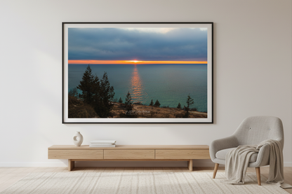 Large fine art landscape print of glowing orange sunset stripe on horizon with golden reflection path across turquoise Lake Michigan viewed from Empire Bluffs overlook with silhouetted evergreens Sleeping Bear Dunes April 2023 displayed in modern living room