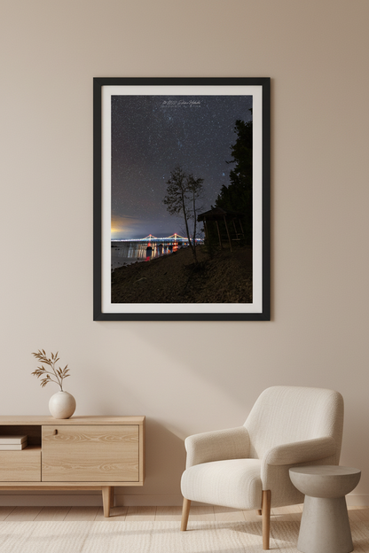 Large fine art portrait print of the Mackinac Bridge illuminated at night with colorful lights reflecting in the Straits of Mackinac silhouetted tree and cabin foreground star filled sky Mackinaw City September 30 2022 displayed in modern living room