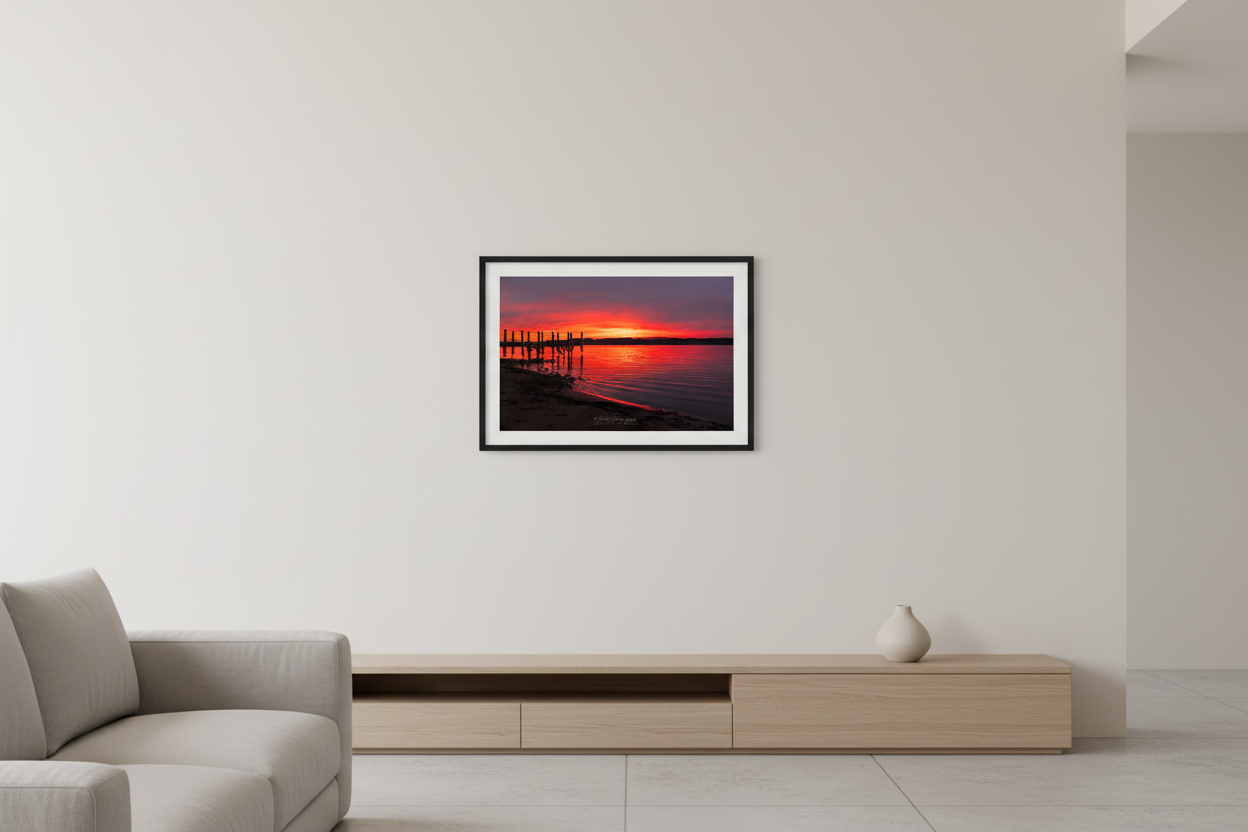 Small fine art print of fiery crimson red orange sunrise over Suttons Bay Leelanau County Michigan with wooden dock pilings silhouette dark treeline reflection in calm water November 3 2024 displayed in modern living room