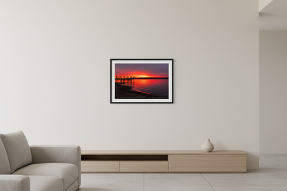 Small fine art print of fiery crimson red orange sunrise over Suttons Bay Leelanau County Michigan with wooden dock pilings silhouette dark treeline reflection in calm water November 3 2024 displayed in modern living room