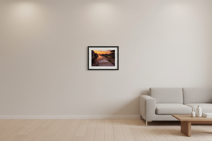 Small fine art long exposure landscape print of Fishtown Leland River channel at sunset with silky smooth water reflecting blazing orange and gold sky charming fishtown shanties docks colorful string lights and boats with open Lake Michigan horizon glowing in distance June 2021 displayed in modern living room