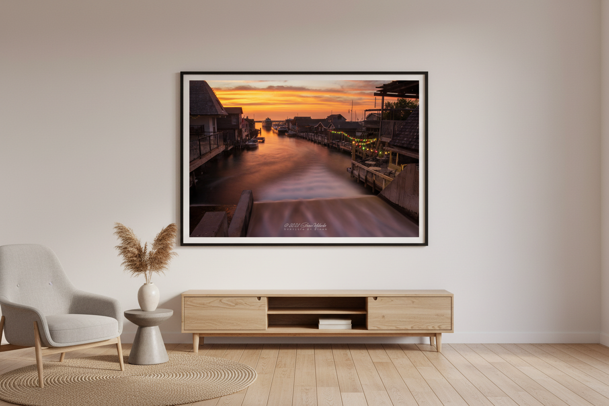 Large fine art long exposure landscape print of Fishtown Leland River channel at sunset with silky smooth water reflecting blazing orange and gold sky charming fishtown shanties docks colorful string lights and boats with open Lake Michigan horizon glowing in distance June 2021 displayed in modern living room