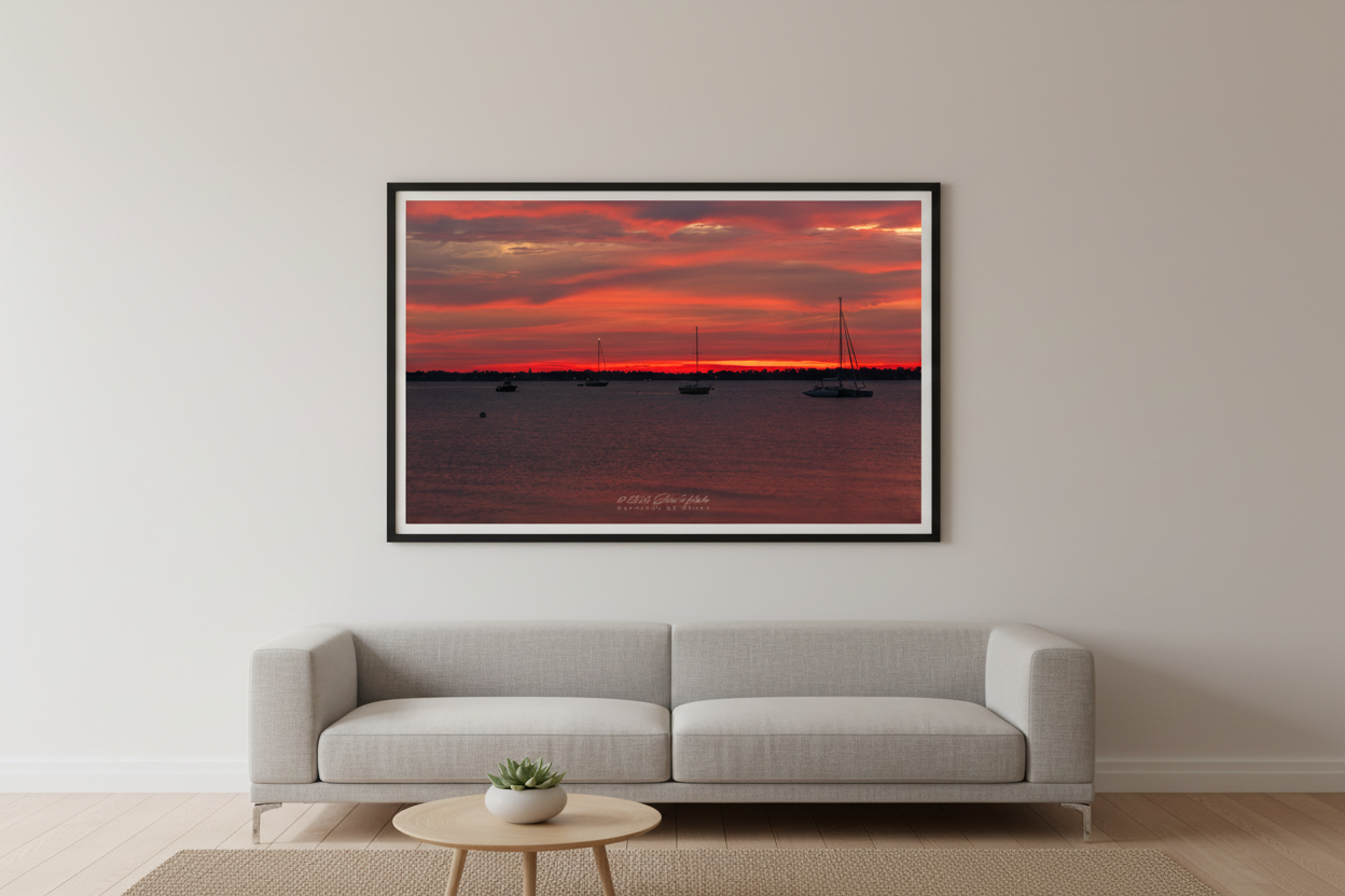 Large fine art print of blazing red fire sunrise over Northport Marina with fleet of sailboat silhouettes against intensely crimson layered sky over dark moody harbor water displayed in modern living room
