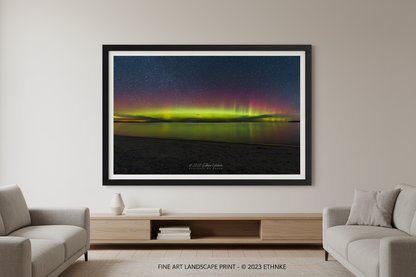 Large fine art landscape print of vivid yellow-green aurora band with crimson red pillars reflecting over Lake Michigan from Glen Haven beach with dark sandy foreground and star-filled sky March 14 2023 displayed in modern living room