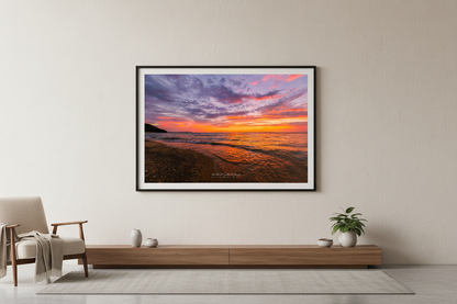 Large fine art landscape print of sweeping wide-angle purple and orange sunset sky with golden wave reflections and rocky shoreline curving toward forested headland at Leland Lake Michigan September 2023 displayed in modern living room