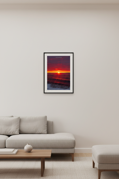 Small fine art portrait print of starburst sunset with deep crimson and purple clouds blazing yellow-orange horizon golden reflection across dark Lake Michigan waves and snow-dusted icy shoreline at Van's Beach Leland Michigan March 2023 displayed in modern living room