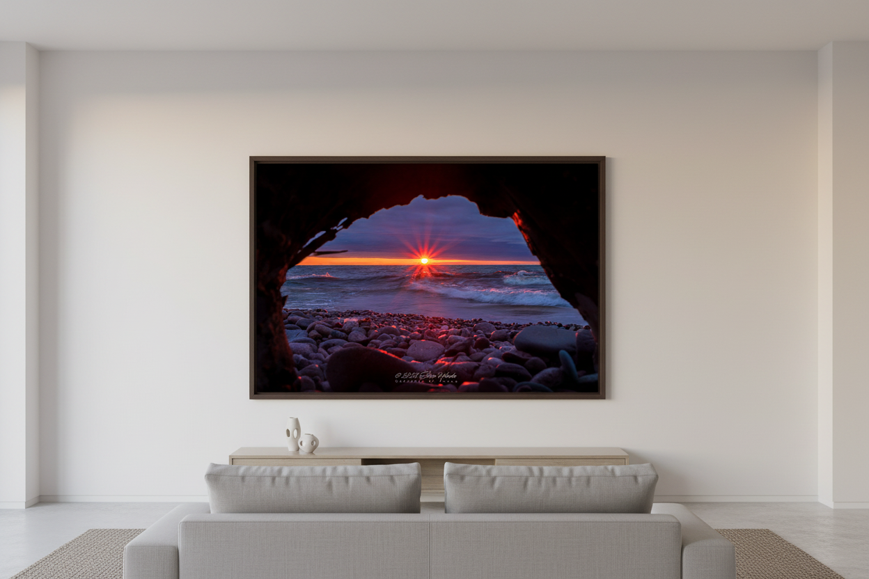 Large fine art landscape print of starburst sunset framed through driftwood rock arch over crashing waves on smooth pebble beach at Eagle River Lake Superior Michigan April 2023 displayed in modern living room