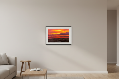 Small fine art print of deep orange red gold sunrise over Sleeping Bear Bay from beach level with flat sandy foreground prominent Sleeping Bear Bluffs silhouette and dark purple cloud mass displayed in modern living room