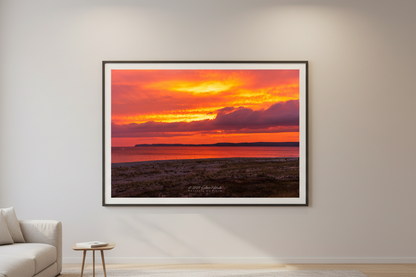 Large fine art print of deep orange red gold sunrise over Sleeping Bear Bay from beach level with flat sandy foreground prominent Sleeping Bear Bluffs silhouette and dark purple cloud mass displayed in modern living room