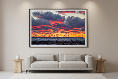 Large fine art landscape print of explosive fiery crimson and magenta layered clouds lit like flames over golden-orange horizon with crashing white-capped waves against rocky breakwall and dark distant headland silhouette at Leland Lake Michigan December 2021 displayed in modern living room