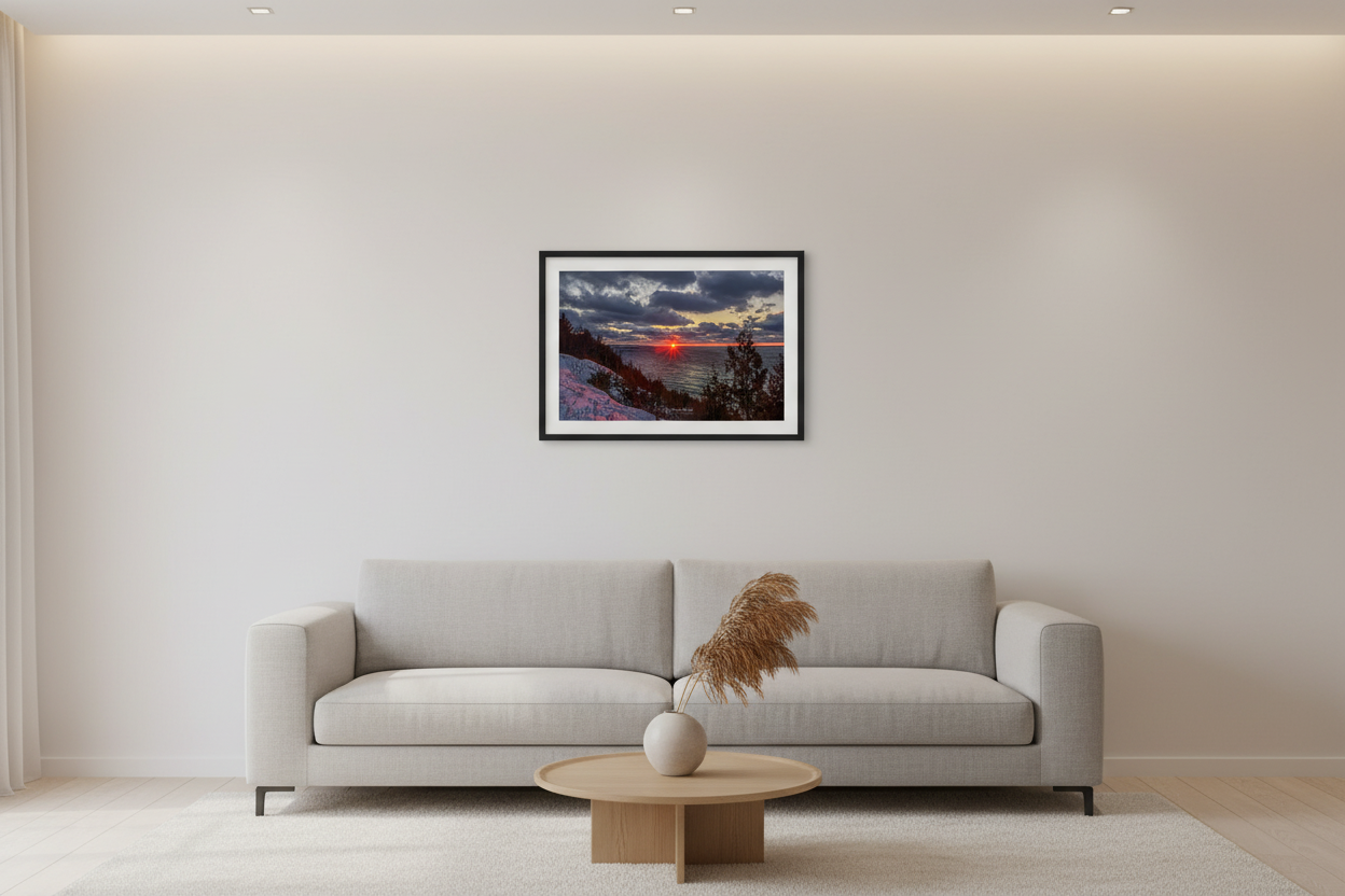 Small fine art landscape print of dramatic starburst sunset with fiery red sun touching Lake Michigan horizon under moody dark storm clouds with golden edges viewed from snow-covered bluff with silhouetted evergreen trees at Sleeping Bear Dunes January 2023 displayed in modern living room