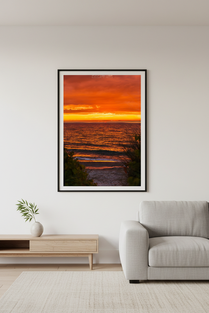 Large fine art portrait print of blazing all-red orange sky sunset with golden wave reflections and lush green foliage beach path framing foreground Leland Lake Michigan July 2024 displayed in modern living room