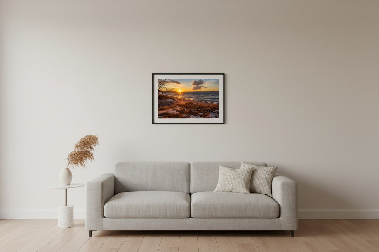 Small fine art landscape print of golden starburst sunset raking warm light across snow-dusted sandy beach with glowing dune grass bare winter trees and rolling Lake Michigan waves at Leland Michigan February 2023 displayed in modern living room