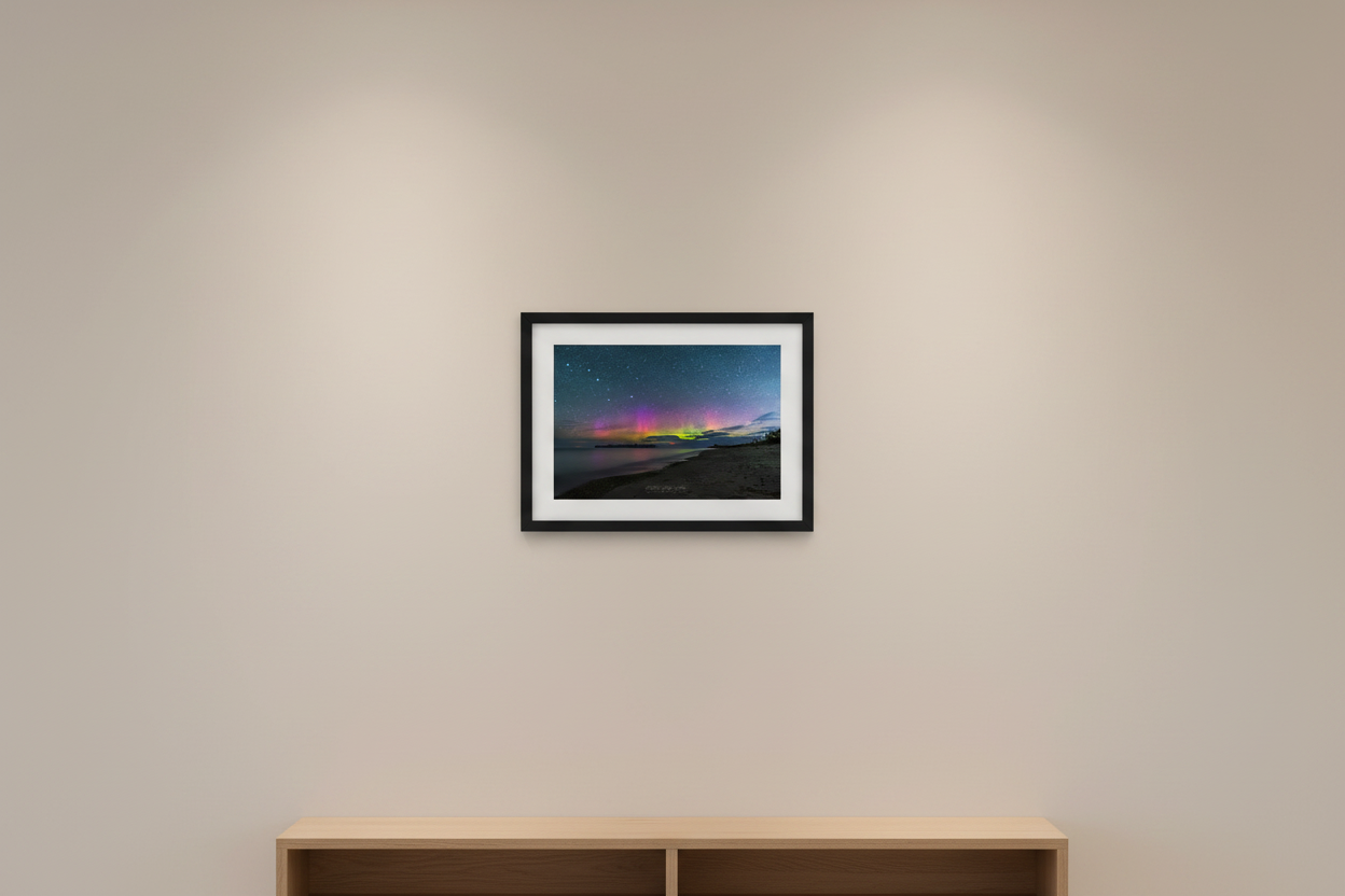 Small fine art landscape print of vivid Northern Lights with magenta purple and neon green aurora bands over glassy calm Lake Michigan shoreline with dark forested peninsula silhouetted on horizon and sandy beach at Fisherman's Island Charlevoix Michigan August 19 2022 displayed in modern living room