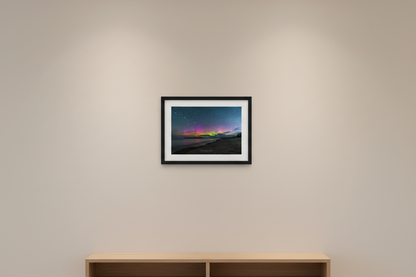 Small fine art landscape print of vivid Northern Lights with magenta purple and neon green aurora bands over glassy calm Lake Michigan shoreline with dark forested peninsula silhouetted on horizon and sandy beach at Fisherman's Island Charlevoix Michigan August 19 2022 displayed in modern living room